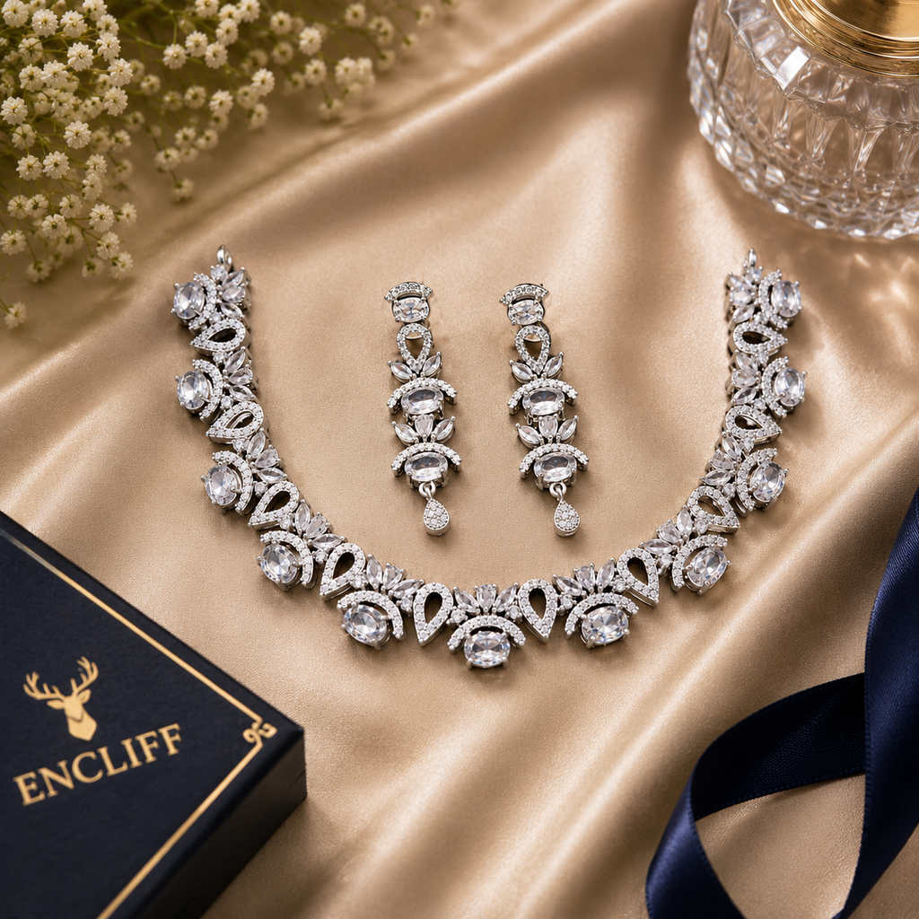 An ornate silver-toned jewelry set with a necklace, matching earrings, and a black ENCLIFF box on a champagne satin backdrop with small white flowers and a glass in the corner.