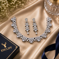 An ornate silver-toned jewelry set with a necklace, matching earrings, and a black ENCLIFF box on a champagne satin backdrop with small white flowers and a glass in the corner.