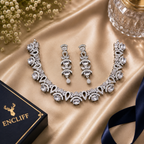 An ornate silver-toned jewelry set with a necklace, matching earrings, and a black ENCLIFF box on a champagne satin backdrop with small white flowers and a glass in the corner.
