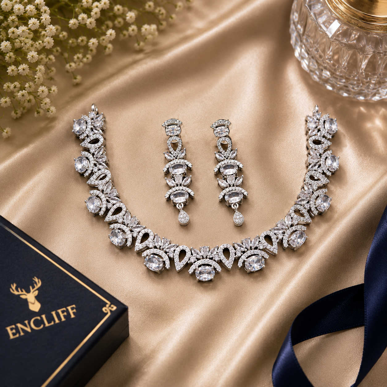 An ornate silver-toned jewelry set with a necklace, matching earrings, and a black ENCLIFF box on a champagne satin backdrop with small white flowers and a glass in the corner.