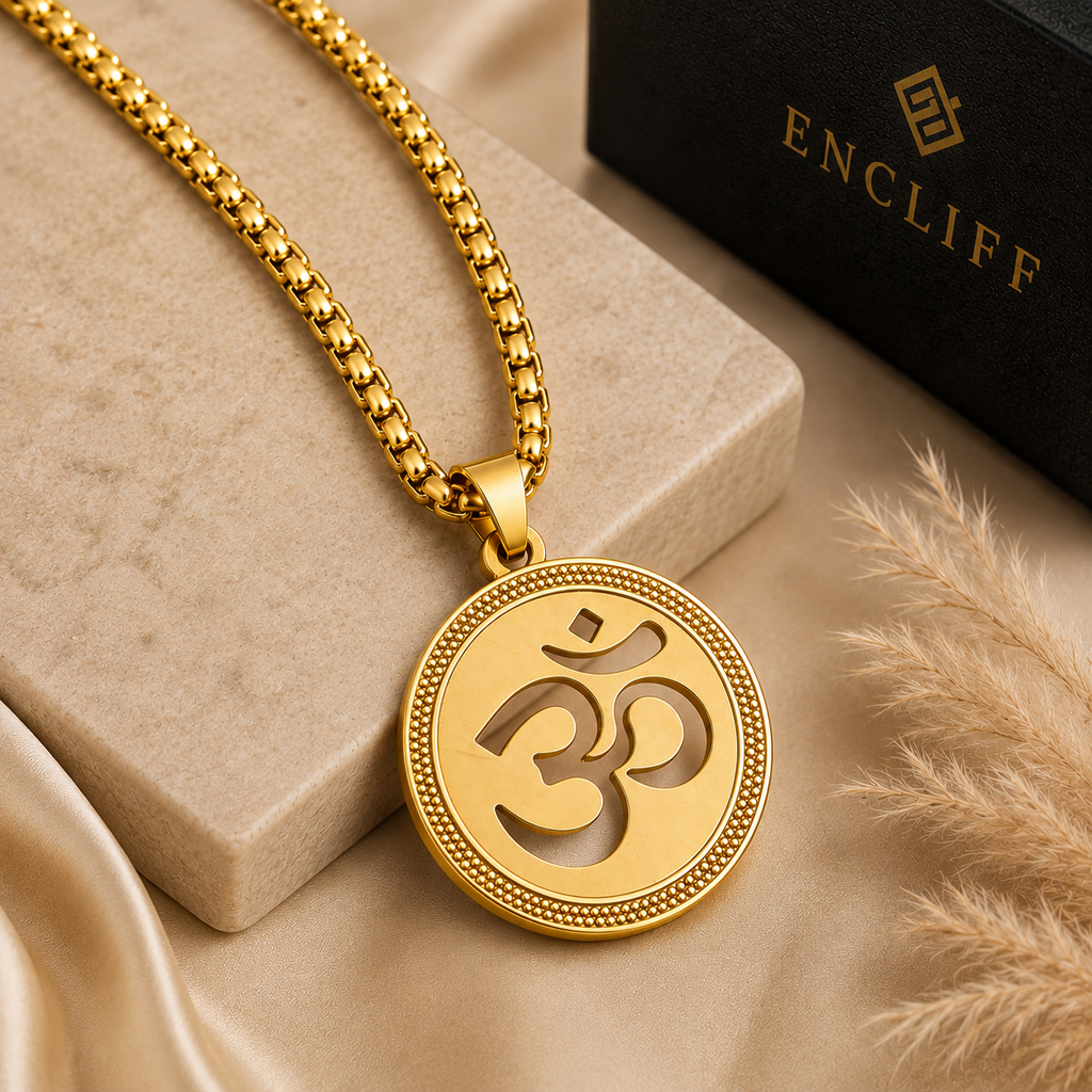 A gold-tone Om pendant necklace on a matching chain, displayed on a beige stone block with a black ENCLIFF box and dried grass in the background.