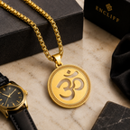 A gold pendant necklace with an Om symbol on a gold chain, displayed on a marble surface with black jewelry boxes, a black watch, and a black cloth. The brand name ENCLIFF is visible on the box in the background.
