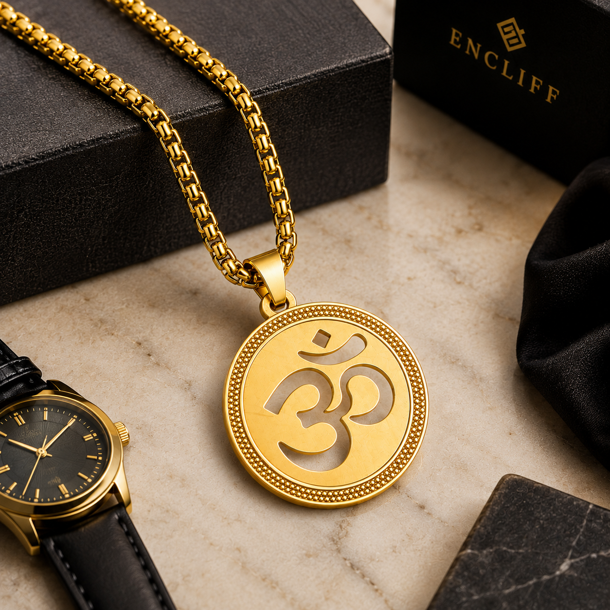 A gold pendant necklace with an Om symbol on a gold chain, displayed on a marble surface with black jewelry boxes, a black watch, and a black cloth. The brand name ENCLIFF is visible on the box in the background.